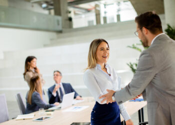 A woman and a man in business attire shake hands and smile in a modern office, possibly after negociating with creditors, while three colleagues talk and work at a table in the background. | MyBestHub.com