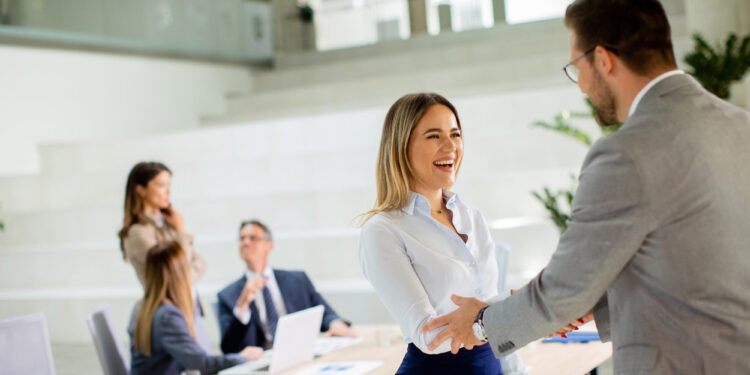 A woman and a man in business attire shake hands and smile in a modern office, possibly after negociating with creditors, while three colleagues talk and work at a table in the background. | MyBestHub.com