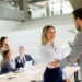 A woman and a man in business attire shake hands and smile in a modern office, possibly after negociating with creditors, while three colleagues talk and work at a table in the background. | MyBestHub.com