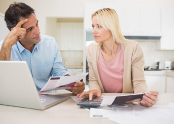 A man and a woman sit at a kitchen table, looking concerned while reviewing documents. The man holds papers, wondering, is debt consolidation a good idea, as the woman uses a calculator and points to a document beside their laptop. | MyBestHub.com