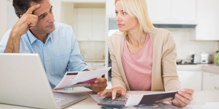 A man and a woman sit at a kitchen table, looking concerned while reviewing documents. The man holds papers, wondering, is debt consolidation a good idea, as the woman uses a calculator and points to a document beside their laptop. | MyBestHub.com