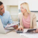 A man and a woman sit at a kitchen table, looking concerned while reviewing documents. The man holds papers, wondering, is debt consolidation a good idea, as the woman uses a calculator and points to a document beside their laptop. | MyBestHub.com