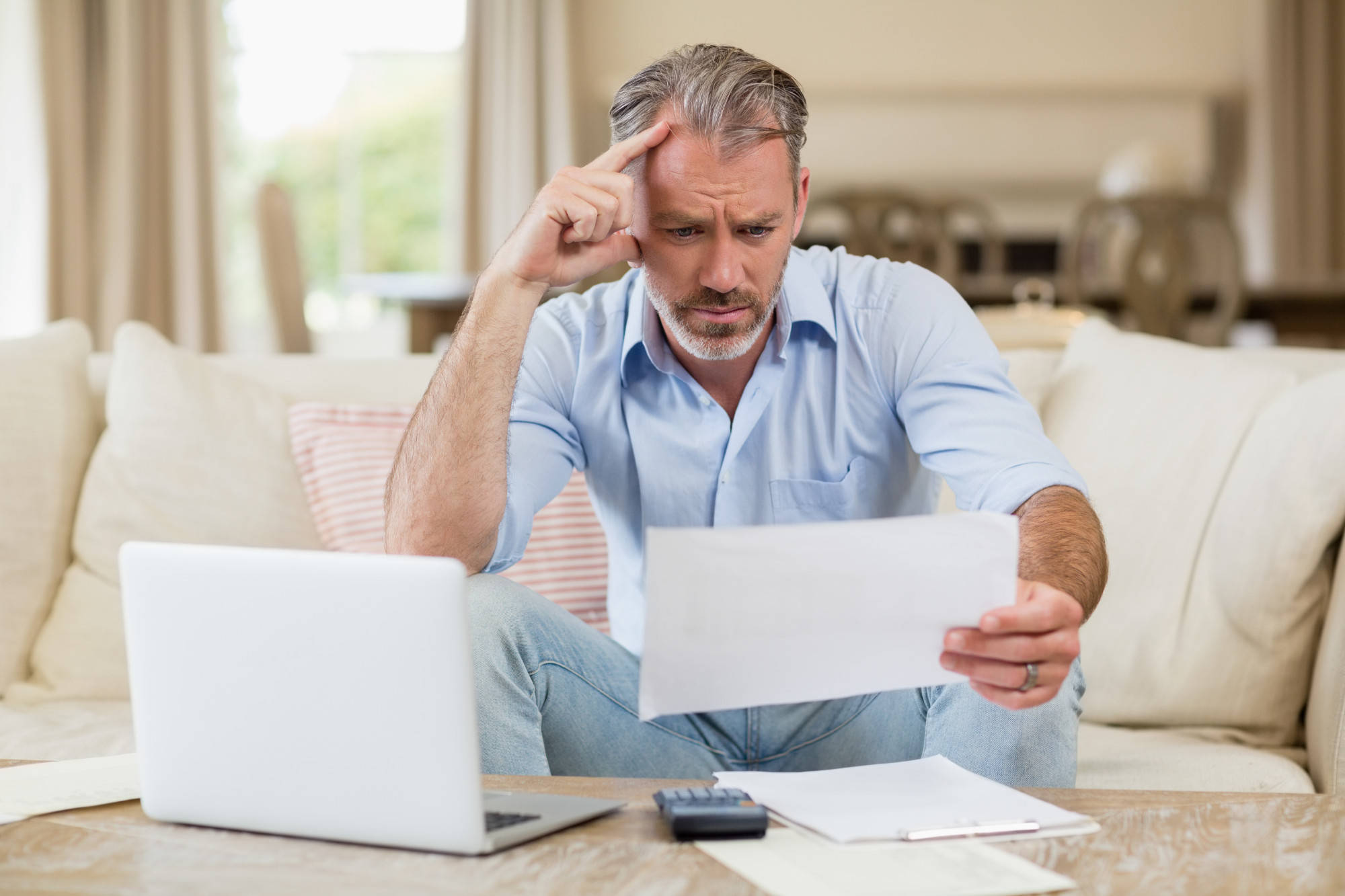 A man sits on a couch, looking concerned as he reads a document. An open laptop, papers, and a calculator on the table suggest he's reviewing finances or bills—perhaps weighing the debt snowball vs avalanche methods for repayment. | MyBestHub.com