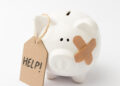 A white ceramic piggy bank with a bandage on its face and a cardboard tag that reads “HELP!” attached to its ear, set against a plain white background. | MyBestHub.com