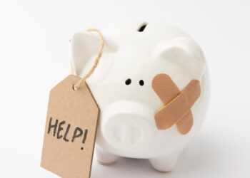 A white ceramic piggy bank with a bandage on its face and a cardboard tag that reads “HELP!” attached to its ear, set against a plain white background. | MyBestHub.com