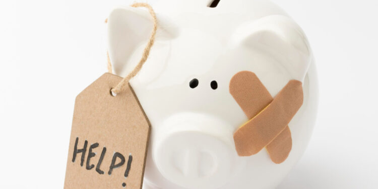 A white ceramic piggy bank with a bandage on its face and a cardboard tag that reads “HELP!” attached to its ear, set against a plain white background. | MyBestHub.com