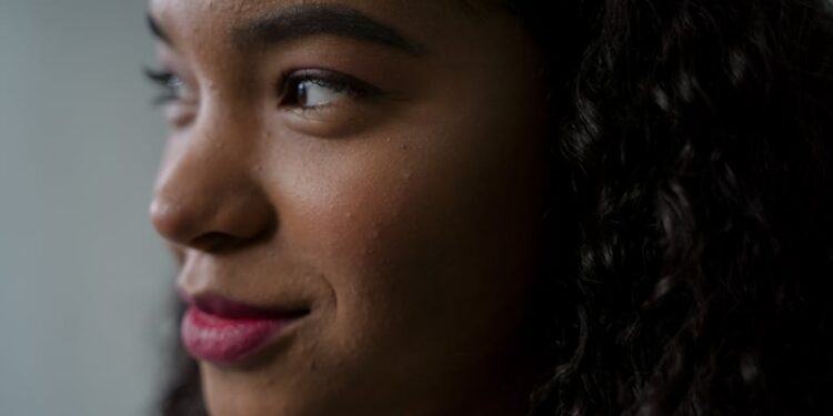 Close-up of a young woman with curly hair looking to the left, showing a thoughtful expression. The background is blurred, focusing attention on her face and natural features. | MyBestHub.com