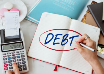 A person writes “DEBT” in a notebook beside a calculator, papers, and a receipt, suggesting financial planning or budgeting. | MyBestHub.com