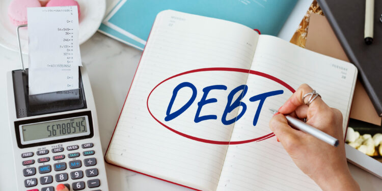 A person writes “DEBT” in a notebook beside a calculator, papers, and a receipt, suggesting financial planning or budgeting. | MyBestHub.com