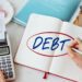 A person writes “DEBT” in a notebook beside a calculator, papers, and a receipt, suggesting financial planning or budgeting. | MyBestHub.com