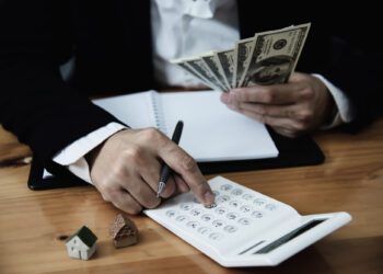 A person in a suit uses a calculator while holding several US dollar bills, with a notebook and small model houses on the wooden table nearby. | MyBestHub.com
