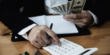 A person in a suit uses a calculator while holding several US dollar bills, with a notebook and small model houses on the wooden table nearby. | MyBestHub.com
