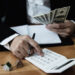 A person in a suit uses a calculator while holding several US dollar bills, with a notebook and small model houses on the wooden table nearby. | MyBestHub.com